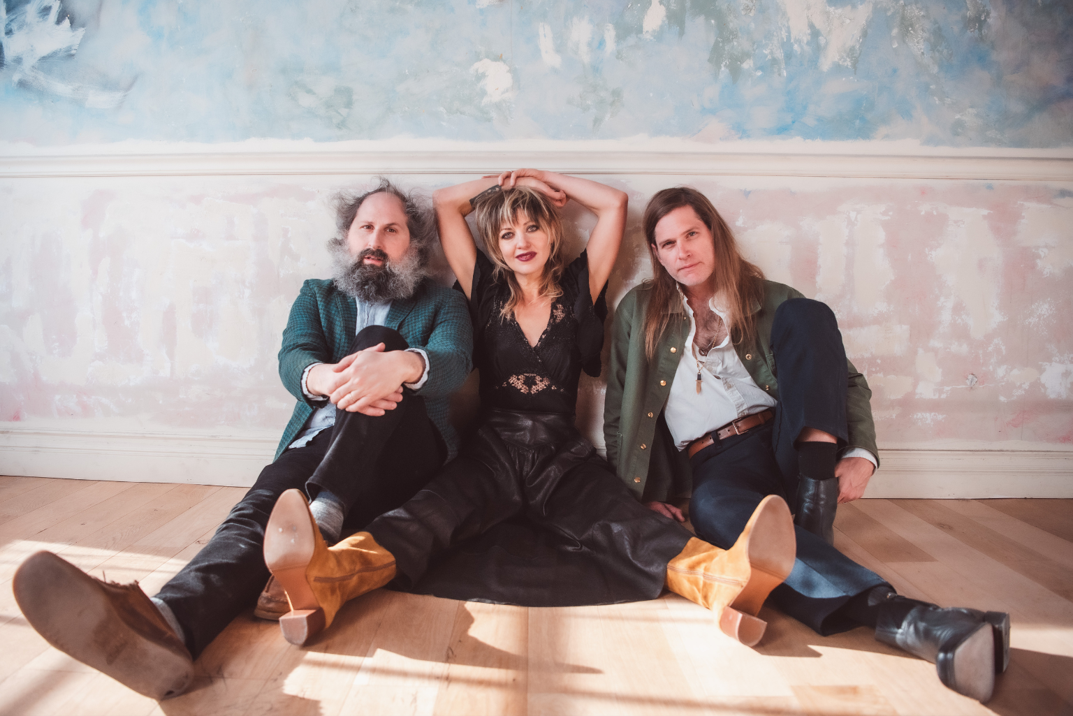 The three musicians sit on the floor against a decorative wall and face the camera.