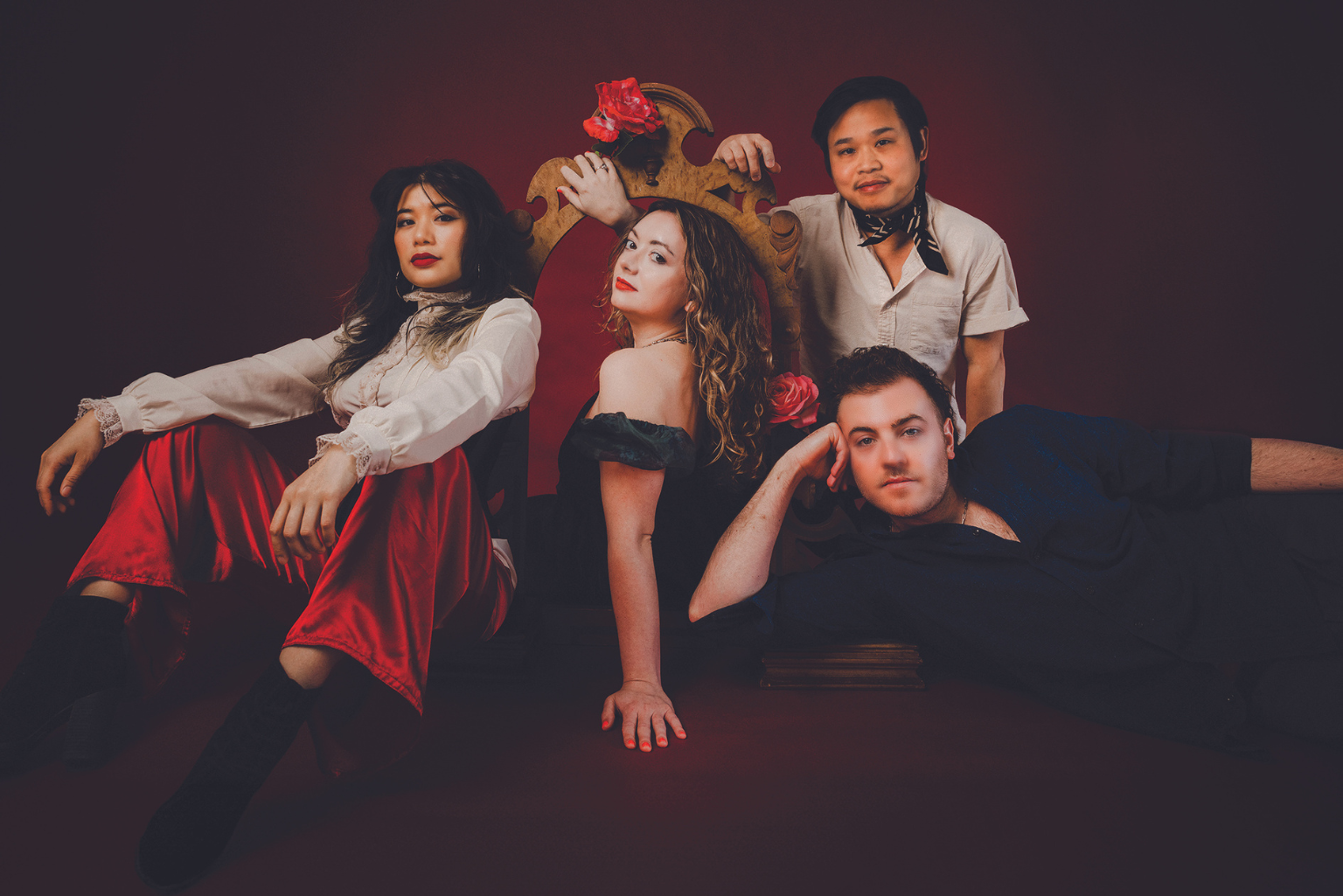 Four musicians seated, crouched, and posing in front of a red backdrop.